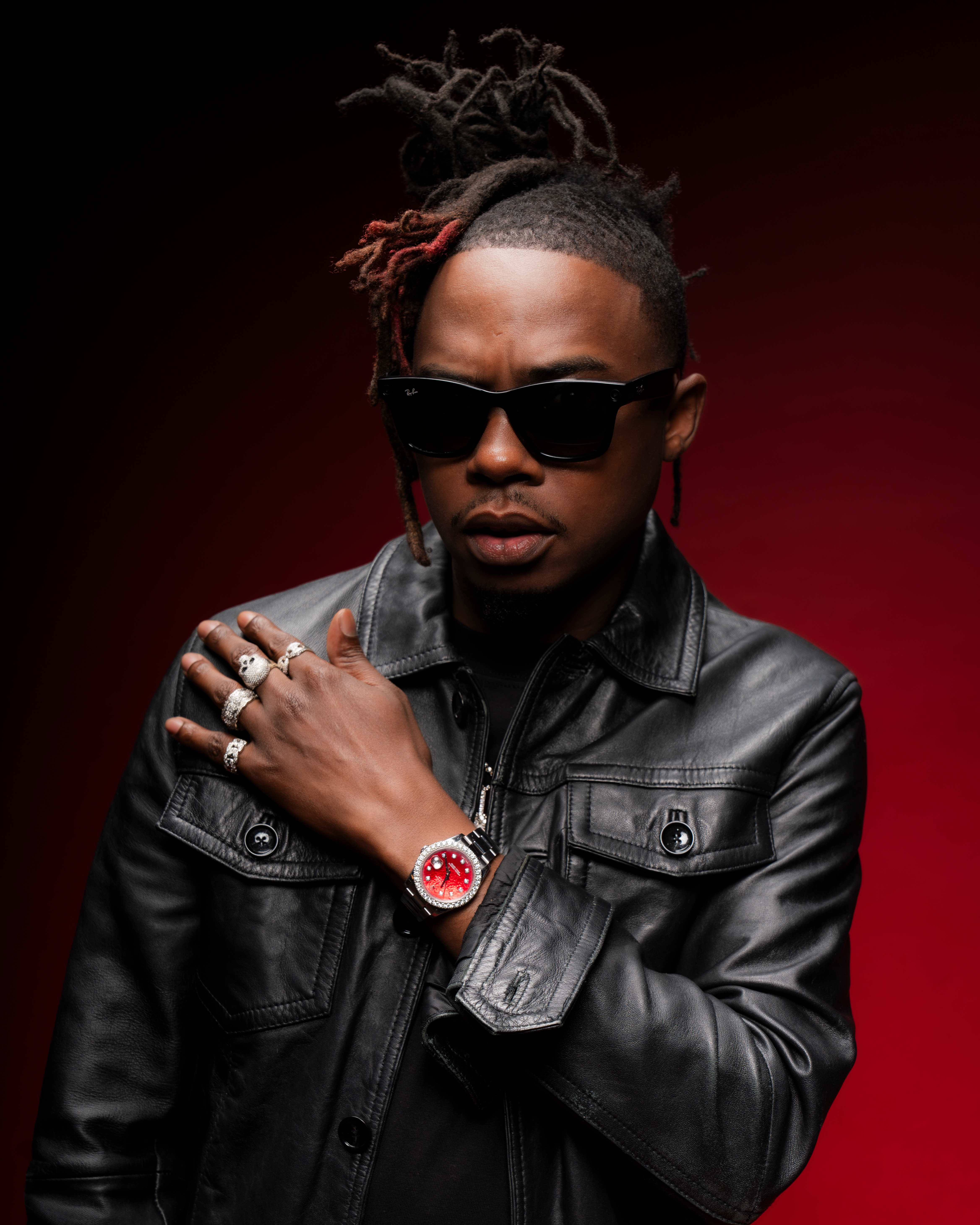 Trap Beckham in black leather with arm crossed over his shoulder against a red backdrop
